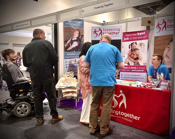 The Fostering Together team at the Disability Expo 2023. The image shows two people looking at our material whilst Neil and Jack from Fostering Together are also chatting to another person at the stand. The stand has lots of information on banners and on the table and the table is coverred with a cloth with Fostering Together's signature pink and the heart logo.