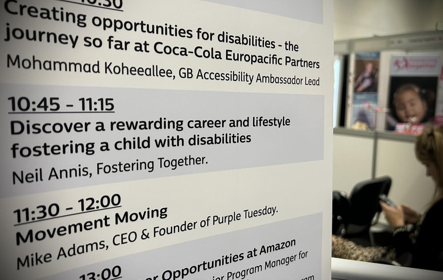 A closeup image of a list of speakers at Disability Expo. In the background is a glimpse of the Fostering Together stand. On the the list above Fostering Together is a speech by Coca Cola 'Creating opportunities for disabilities' and following us is the CEO of Purple Thursday, followed by a talk by Amazon talking about their career opportunities for people who have disabilties. Fostering Together's slot is 'Discover a rewarding career and lifestyle fostering a child with disabilities' presented by Neil Annis.