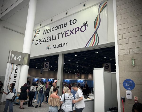 A view of the crowds at the entrance of the event. A large banner is above the entrance saying 'Welcome to Disability EXPO'. Many groups of people are standing and reviewing the event programmes.